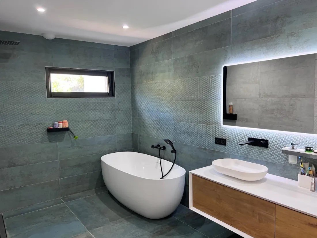 Salle de bain carrelée effet béton avec baignoire îlot, vasque et miroir rétroéclairé