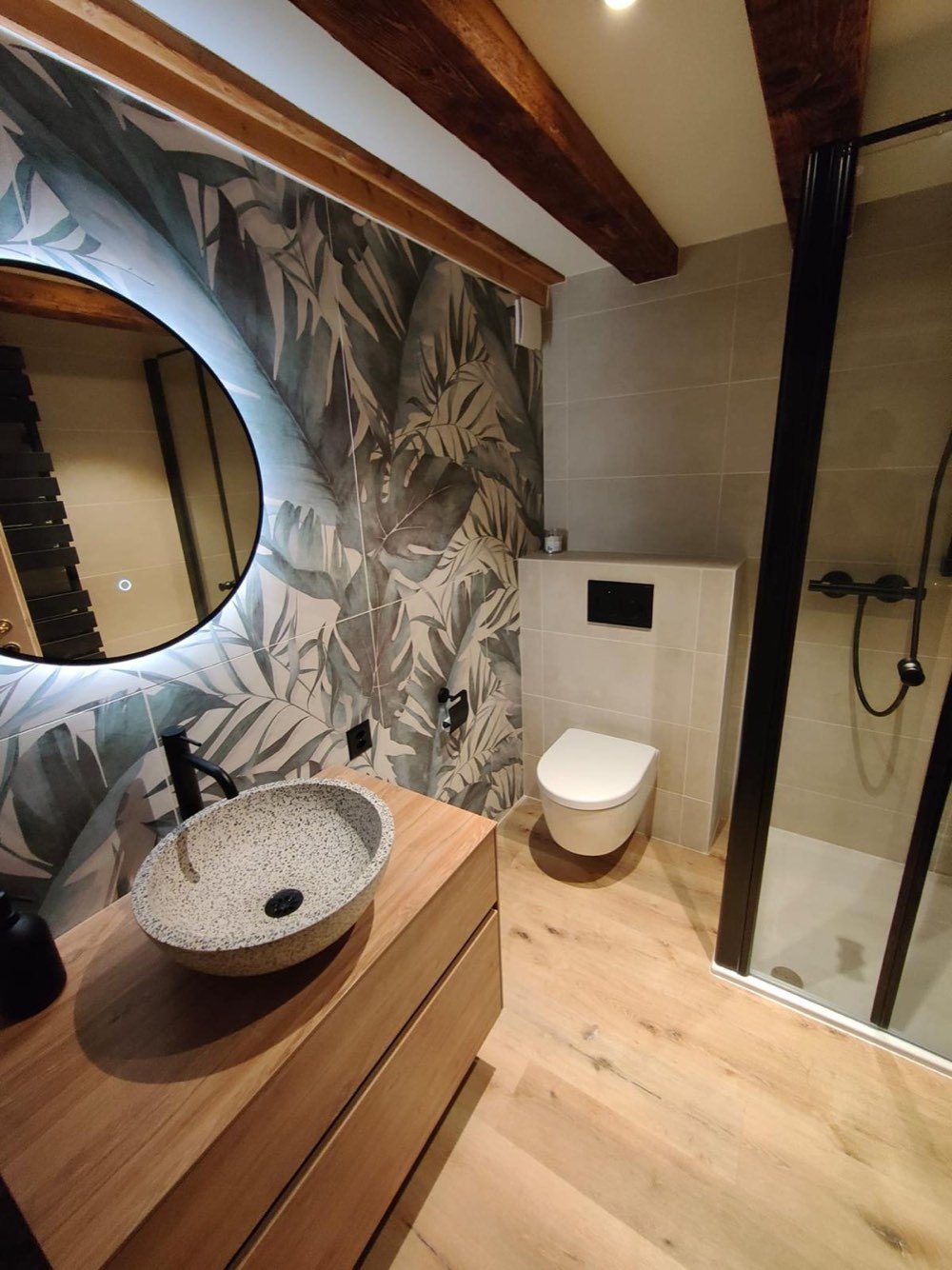 Salle de bain rénovée sous combles : douche vitrée, WC suspendu et vasque en terrazzo sur meuble en chêne, poutres apparentes